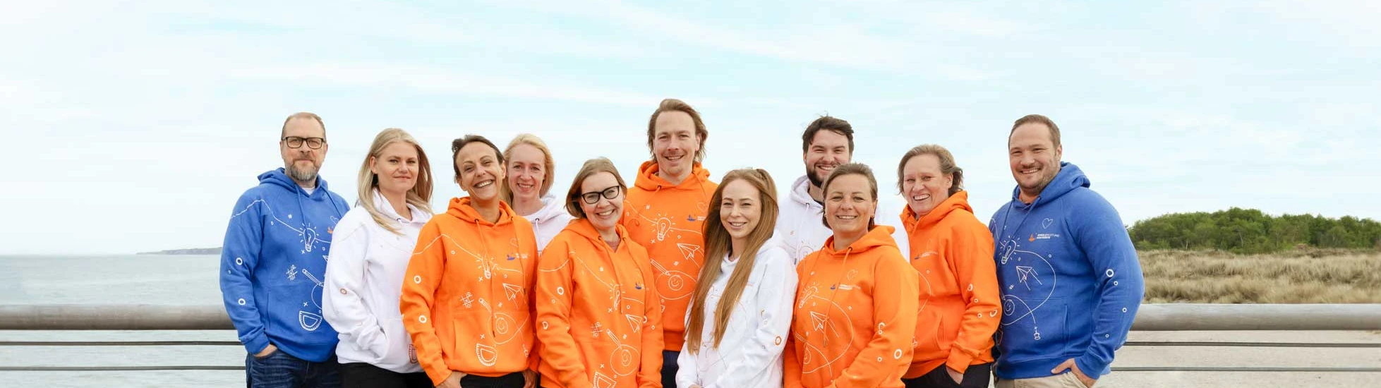 Gruppenfoto des Teams von Schulbegleitung Westküste auf einer Promenade am Strand mit den neuen Kapuzenpullovern in blau, weiß und orange.