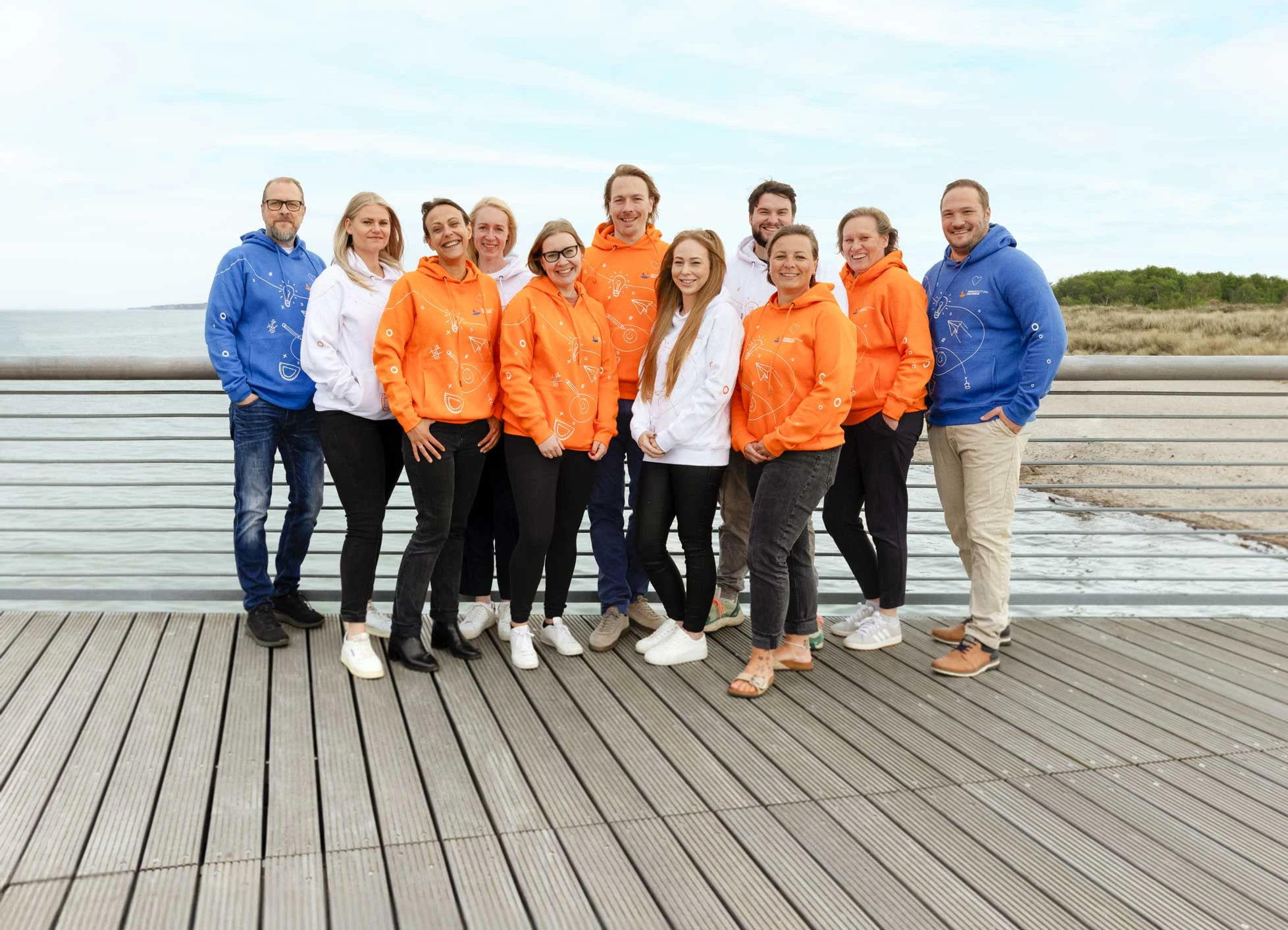 Gruppenfoto des Teams von Schulbegleitung Westküste auf einer Promenade am Strand mit den neuen Kapuzenpullovern in blau, weiß und orange.