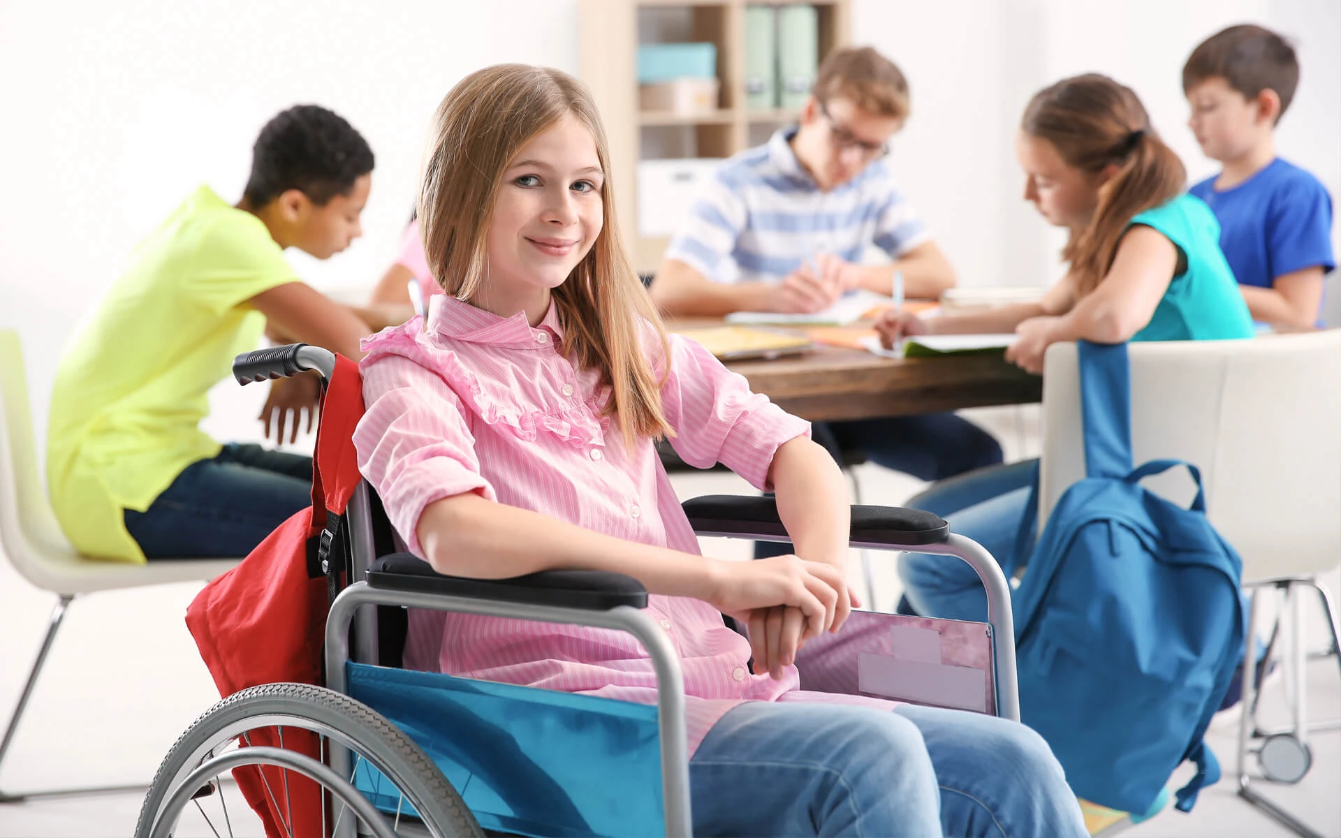 Ein Mädchen in einem rosa Pullover sitzt mit vier weiteren Kindern in einem Klassenraum. Sie sitzt im Rollstuhl.