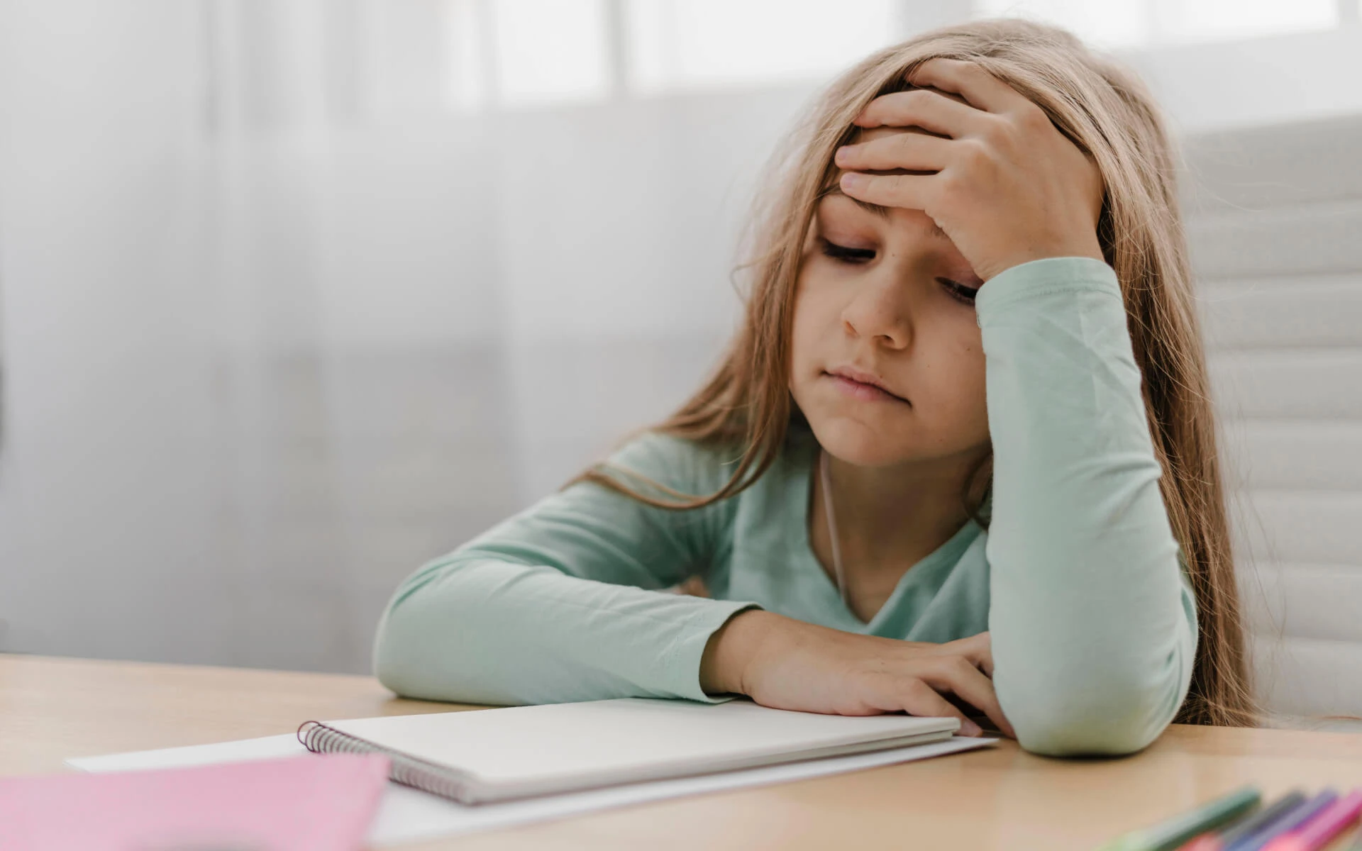 Ein Kind sitzt mit einem leeren Block an einem Tisch. Es hält eine Hand an die Stirn und sieht traurig aus.